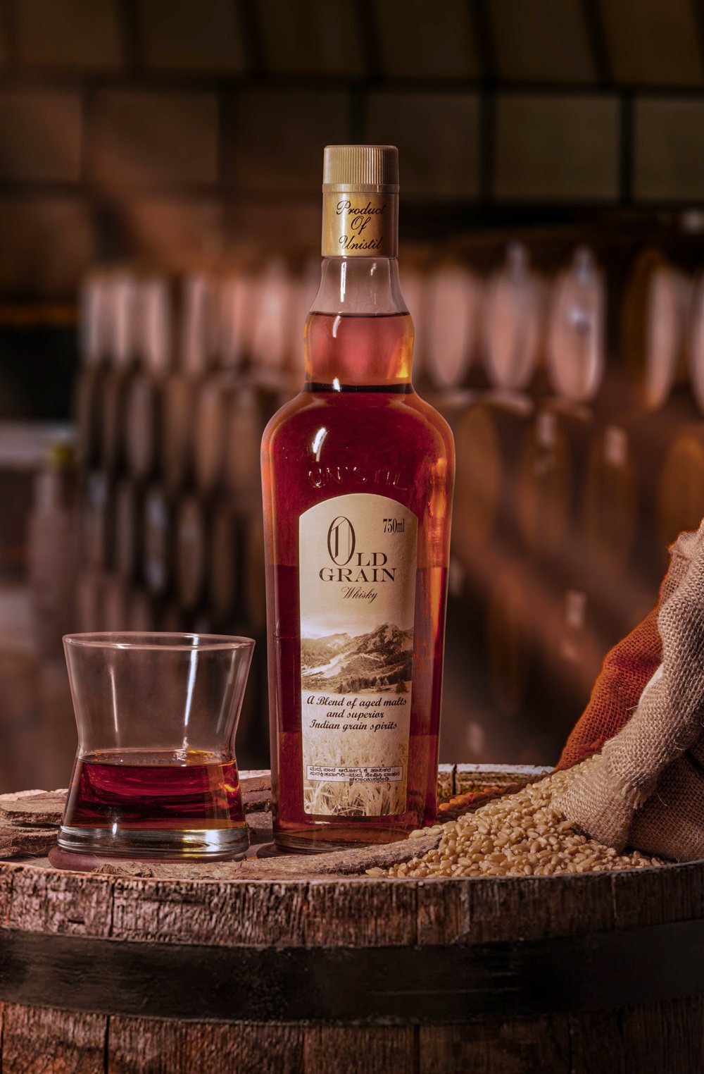 A bottle of Old Grain Whisky by Unistil on a barrel, with a glass and a sack of grain